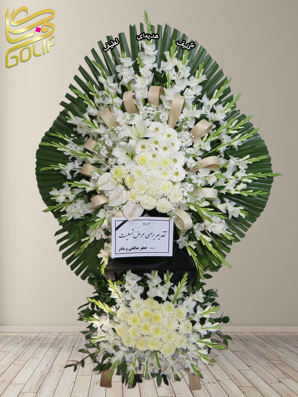 تاج گل تسلیت یک و نیم طبقه (لیلیوم، گلایل، داوودی) گل‌های اصلی و غالب این تاج گل شامل موارد زیر هستند: لیلیوم‌های سفید: این گل‌ها برجسته‌ترین نقش را در ترکیب دارند و در بخش‌های مختلف تاج گل مشاهده می‌شوند. گلایل‌های سفید: که به زیبایی در طراحی تاج گل به‌خصوص در نقاط بالا و مرکزی ظاهر شده‌اند. داوودی‌های سفید: در نقاط پایین‌تر ترکیب حضور دارند و جلوه‌ای آرامش‌بخش و ملایم به تاج گل می‌دهند. این تاج گل که برای مراسم ختم و ترحیم و تسلیت تقدیم شده، نمادی از احترام و بزرگداشت است. تاج گل ختم، تاج گل ترحیم، تاج گل مراسم ترحیم سفارش آنلاین تاج گل سفارش آنلاین گل عکاسی شده توسط گل‌فروشی آنلاین گلیف