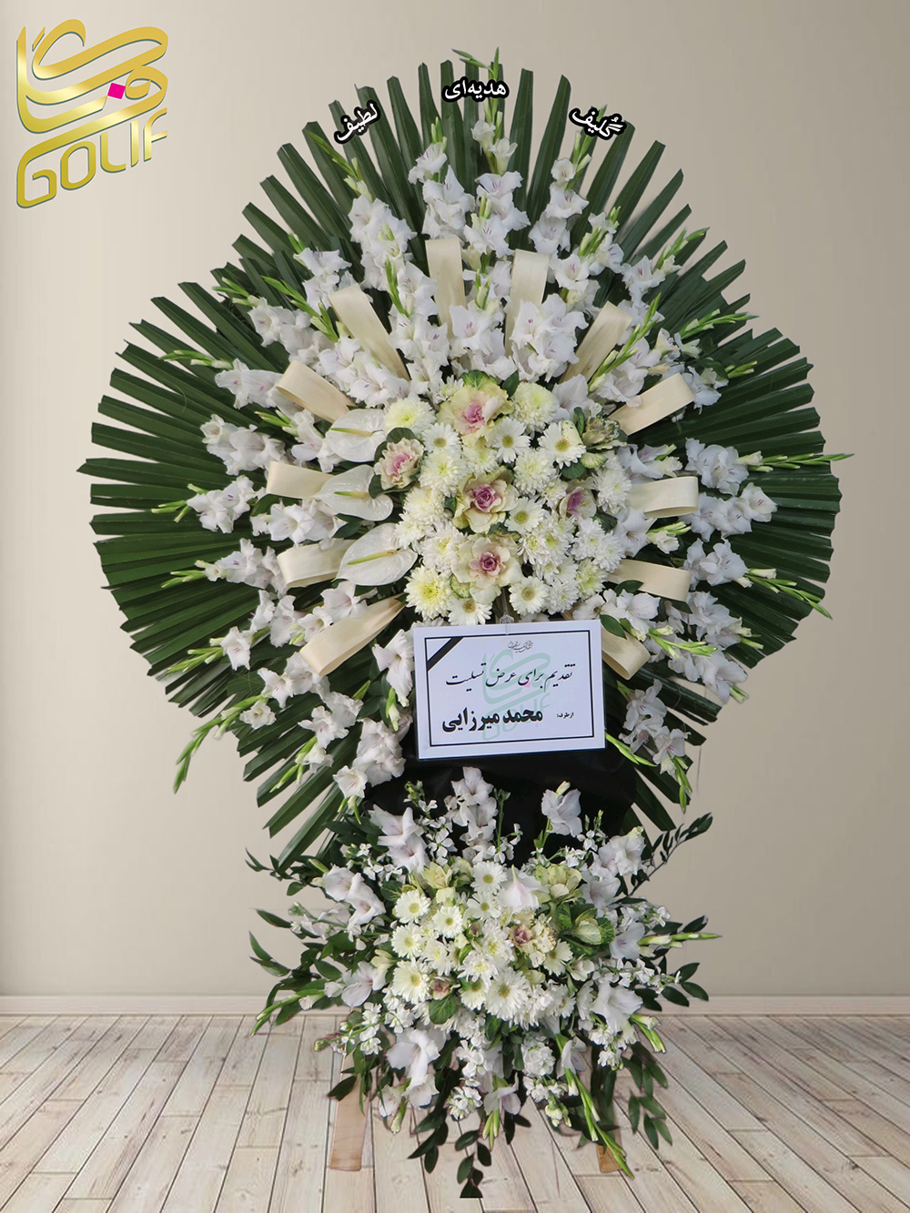 تاج گل تسلیت یک و نیم طبقه (لیلیوم، گلایل، داوودی) تاج گل ختم، تاج گل ترحیم، تاج گل مراسم ترحیم لیلیوم‌های سفید: که در مرکز و بخش‌هایی از طراحی اصلی نمای برجسته‌ای دارند. گلایل‌های سفید: به عنوان یکی دیگر از گل‌های اصلی استفاده شده و در اطراف تاج گل قرار دارند. داوودی‌های سفید: به صورت پراکنده در کل طراحی دیده می‌شوند و جلوه‌ای آرام و دلنشین ایجاد کرده‌اند. همچنین، استفاده از برگ‌های سبز در پس‌زمینه به تعادل و هماهنگی کلی طراحی کمک کرده است. این تاج گل با پیام تسلیت روی کارت، سمبلی از همدردی و احترام عمیق است. سفارش آنلاین تاج گل سفارش آنلاین گل عکاسی شده توسط گل‌فروشی آنلاین گلیف